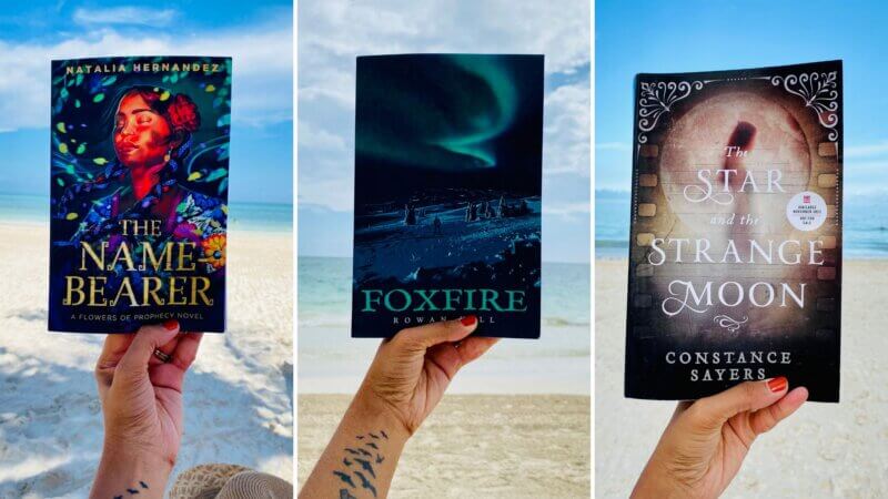 a left hand with a bird tattoo holding three books with the ocean and white beach sand in the background. - the name-bearer, foxfire and The star and the strange moon.