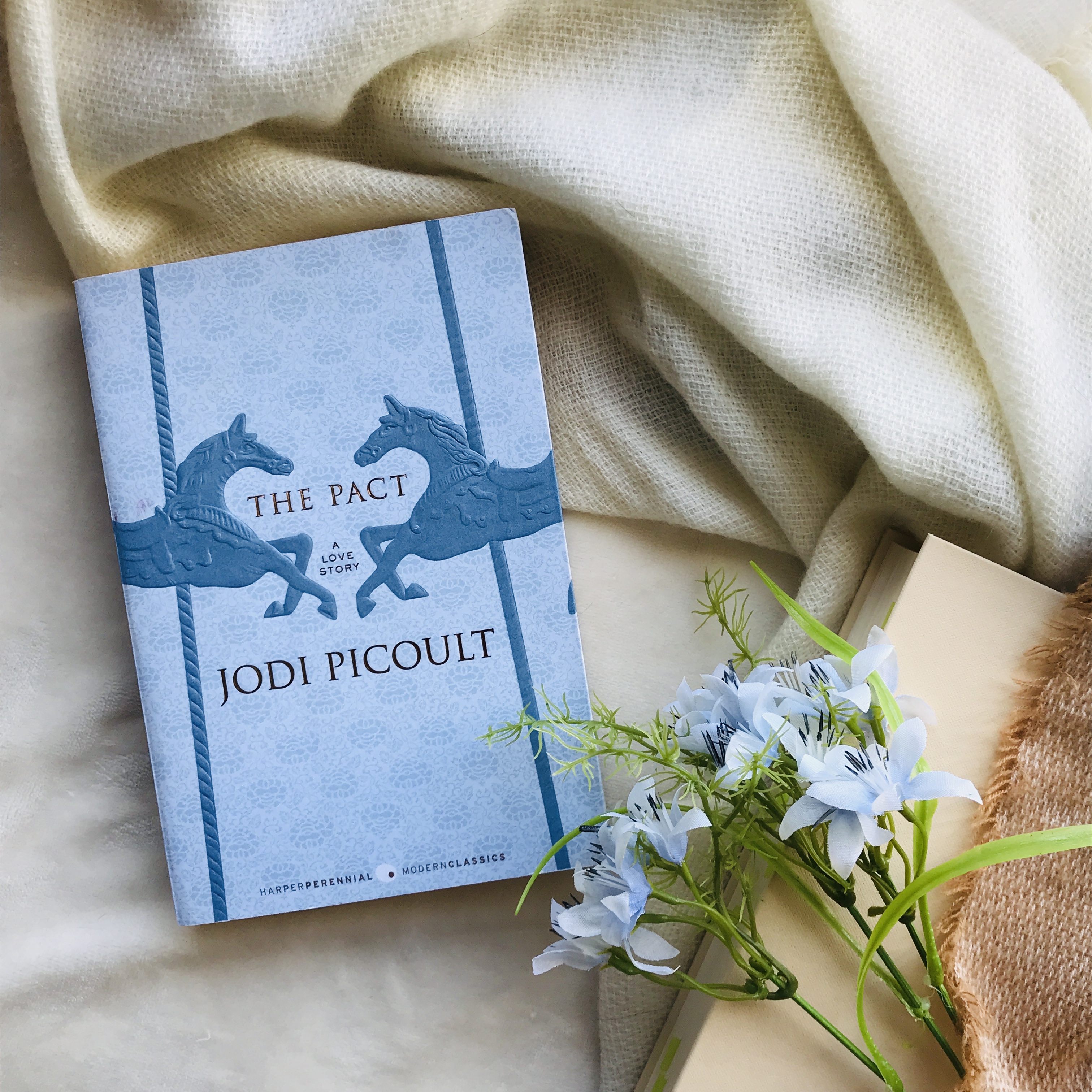 My copy of The Pact by Jodi Picoult close to blue flowers and a blanket